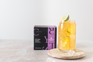 A glass filled with a refreshing beverage containing ice, lemon, and lime slices is placed on a wooden coaster beside a black and purple box labeled 'Elixir Detox AGATA' from Crystaltea. The setup is against a light, minimalist background.