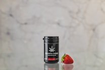 A small black container labeled 'CRYSTALWEED HIGH QUALITY CBD' is placed next to a ripe, red strawberry. The surfaces reflect on the shiny tabletop, and the background is a neutral, blurry marble texture.