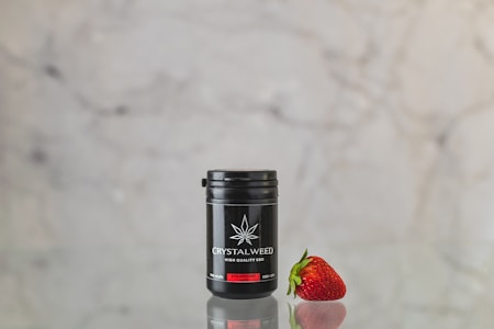A small black container labeled 'CRYSTALWEED HIGH QUALITY CBD' is placed next to a ripe, red strawberry. The surfaces reflect on the shiny tabletop, and the background is a neutral, blurry marble texture.