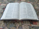 An open bilingual book featuring pages with text in both English and Korean. It is placed on a colorful, patterned fabric surface, possibly a tapestry or carpet.