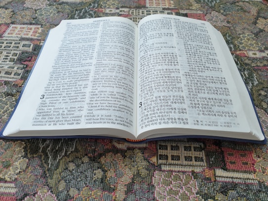 An open bilingual book featuring pages with text in both English and Korean. It is placed on a colorful, patterned fabric surface, possibly a tapestry or carpet.