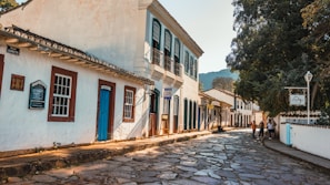 A picturesque cobblestone street lined with colonial-style buildings featuring wooden doors and windows. The structures have white facades with brightly colored trims. There are a few people walking and talking on the sidewalk, and a large tree casts shade over part of the street. Signage in Portuguese indicates this is a historic area.