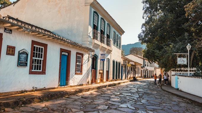 A picturesque cobblestone street lined with colonial-style buildings featuring wooden doors and windows. The structures have white facades with brightly colored trims. There are a few people walking and talking on the sidewalk, and a large tree casts shade over part of the street. Signage in Portuguese indicates this is a historic area.