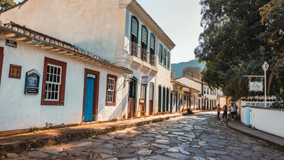 A picturesque cobblestone street lined with colonial-style buildings featuring wooden doors and windows. The structures have white facades with brightly colored trims. There are a few people walking and talking on the sidewalk, and a large tree casts shade over part of the street. Signage in Portuguese indicates this is a historic area.