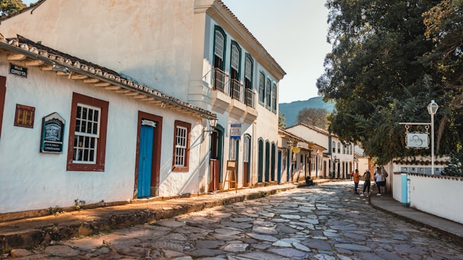A picturesque cobblestone street lined with colonial-style buildings featuring wooden doors and windows. The structures have white facades with brightly colored trims. There are a few people walking and talking on the sidewalk, and a large tree casts shade over part of the street. Signage in Portuguese indicates this is a historic area.