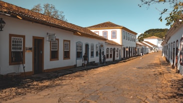 A cobblestone street lined with historic buildings featuring white walls and brown wooden frames. The architecture is colonial, with tiled roofs and decorative window frames. A few people are walking or standing along the street, which adds a sense of leisurely pace. The scene is bathed in warm sunlight and there are trees visible in the background, adding greenery to the urban environment.