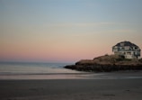 A large, blue house is situated on a rocky outcrop by the ocean. The beach in the foreground is sandy and the sea is calm. The sky is a gradient of soft pink and blue, suggesting either sunrise or sunset.