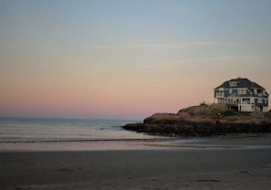 A large, blue house is situated on a rocky outcrop by the ocean. The beach in the foreground is sandy and the sea is calm. The sky is a gradient of soft pink and blue, suggesting either sunrise or sunset.