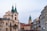 The image displays a street view of a European city with historic architecture. Prominent buildings with steeples and towers frame the scene, and there is a crowd of people visible on the street. Trees without leaves are also noticeable, suggesting a colder season.