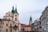The image displays a street view of a European city with historic architecture. Prominent buildings with steeples and towers frame the scene, and there is a crowd of people visible on the street. Trees without leaves are also noticeable, suggesting a colder season.