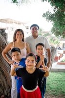 A family of five is posing together outdoors in a park-like setting. The parents stand behind, smiling, with three children in front of them. One child is making hand gestures, and another is wearing glasses and a red skirt. In the background, there are trees and a partially constructed building.