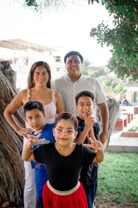 A family of five is posing together outdoors in a park-like setting. The parents stand behind, smiling, with three children in front of them. One child is making hand gestures, and another is wearing glasses and a red skirt. In the background, there are trees and a partially constructed building.