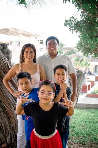 A family of five is posing together outdoors in a park-like setting. The parents stand behind, smiling, with three children in front of them. One child is making hand gestures, and another is wearing glasses and a red skirt. In the background, there are trees and a partially constructed building.