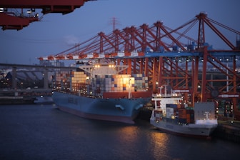 A busy port with containers being loaded onto a cargo ship at sunset.