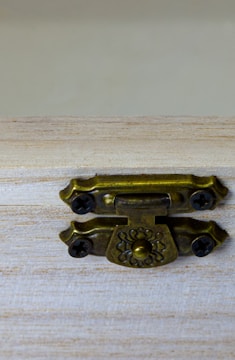 A close-up view of an ornate, metallic latch on a wooden surface. The latch features a decorative design and is fastened with four screws. The wood appears light-colored and slightly textured.