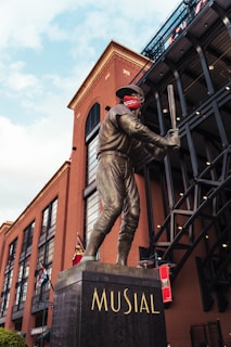 A bronze statue of a baseball player holding a bat is prominently displayed. The statue is mounted on a pedestal with the name 'MUSIAL' inscribed on it. The background features a large brick building with several arched windows and a black metal structure. There are flags hanging from the building, and the sky is partly cloudy.