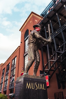 A bronze statue of a baseball player holding a bat is prominently displayed. The statue is mounted on a pedestal with the name 'MUSIAL' inscribed on it. The background features a large brick building with several arched windows and a black metal structure. There are flags hanging from the building, and the sky is partly cloudy.