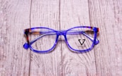 A pair of stylish eyeglasses with blue and brown tortoiseshell frames resting on a textured wooden surface. The lenses are clear, and the brand name 'Veltom' is visible in black on the lens surface.