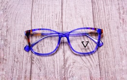 A pair of stylish eyeglasses with blue and brown tortoiseshell frames resting on a textured wooden surface. The lenses are clear, and the brand name 'Veltom' is visible in black on the lens surface.