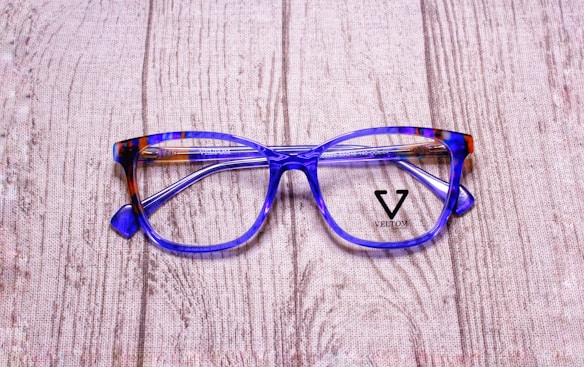 A pair of stylish eyeglasses with blue and brown tortoiseshell frames resting on a textured wooden surface. The lenses are clear, and the brand name 'Veltom' is visible in black on the lens surface.