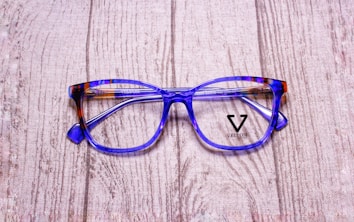 A pair of stylish eyeglasses with blue and brown tortoiseshell frames resting on a textured wooden surface. The lenses are clear, and the brand name 'Veltom' is visible in black on the lens surface.