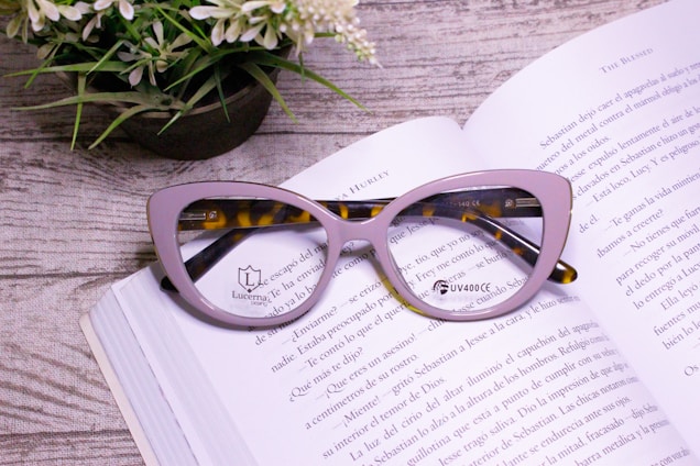 A pair of stylish tortoiseshell and lavender-colored cat-eye glasses rests on an open book with text visible in Spanish. A small potted plant with green leaves and white flowers is positioned next to the book on a wooden surface.