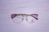 A pair of eyeglasses with a metallic gold frame and pink accents rests on a textured, white wooden surface. The lenses are clear, and there is a logo on the left lens with the word 'VELTOM'.