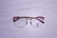 A pair of eyeglasses with a metallic gold frame and pink accents rests on a textured, white wooden surface. The lenses are clear, and there is a logo on the left lens with the word 'VELTOM'.