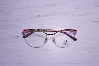 A pair of eyeglasses with a metallic gold frame and pink accents rests on a textured, white wooden surface. The lenses are clear, and there is a logo on the left lens with the word 'VELTOM'.