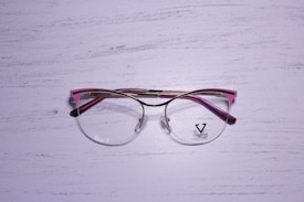 A pair of eyeglasses with a metallic gold frame and pink accents rests on a textured, white wooden surface. The lenses are clear, and there is a logo on the left lens with the word 'VELTOM'.
