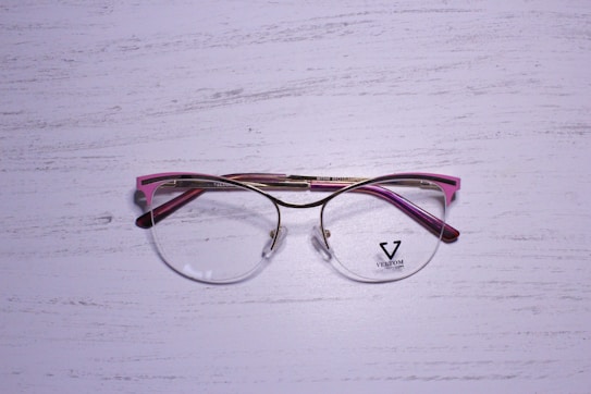 A pair of eyeglasses with a metallic gold frame and pink accents rests on a textured, white wooden surface. The lenses are clear, and there is a logo on the left lens with the word 'VELTOM'.