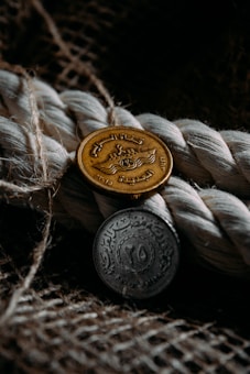 Two intricately designed coins are placed against a textured background of coiled rope and woven fabric. One coin is gold-colored, while the other is silver-toned, each featuring detailed inscriptions and symbols.