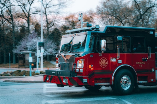 A red fire truck is seen driving through a street intersection. The truck is prominently marked with 'Engine 14' on its side. In the background, there are blurred trees and some street infrastructure, suggesting the vehicle is in motion.