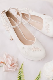 Elegant white bridal shoes adorned with delicate pearl and floral embellishments are placed on a soft, light background. Surrounding the shoes are pale pink roses and green ferns, creating a romantic and refined arrangement.