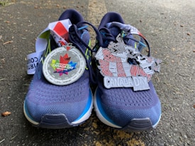 A pair of navy blue running shoes is placed on an asphalt surface. Two medals are resting on the shoes. One medal features a colorful maple leaf design with the text 'Course Virtuelle Canada Day Virtual Run.' The other medal is shaped like a map of Canada with provinces labeled and 'Canada Day' written across it.
