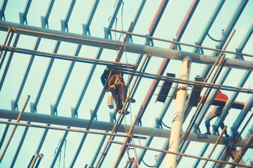 Workers wearing safety gear are engaged in construction activities on a high scaffolding structure. The metal beams and supports form a grid pattern against the sky, with two workers visible, one handling equipment and the other moving along the beams.