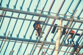 Workers wearing safety gear are engaged in construction activities on a high scaffolding structure. The metal beams and supports form a grid pattern against the sky, with two workers visible, one handling equipment and the other moving along the beams.