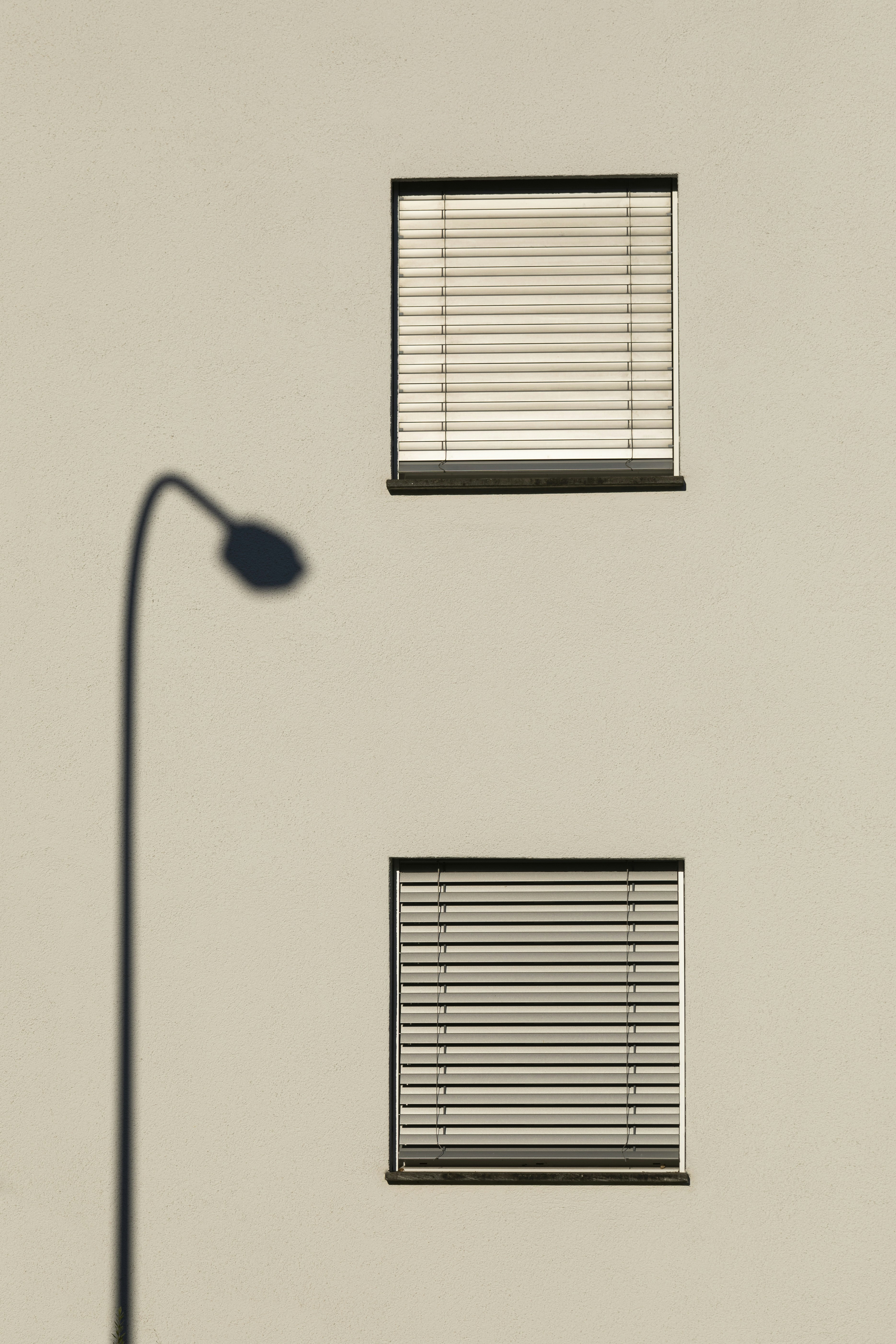 Light-colored exterior wall with two rectangular windows fitted with horizontal blinds and dark trim, alongside the shadow of a street lamp.