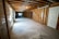 A spacious unfinished basement with exposed wooden beams on the ceiling and concrete floors. The walls are partially covered with wooden framing and plastic sheets. There is a door on the right side and a small window at the end of the room, providing some natural light. Pipes and ductwork are visible along the ceiling.