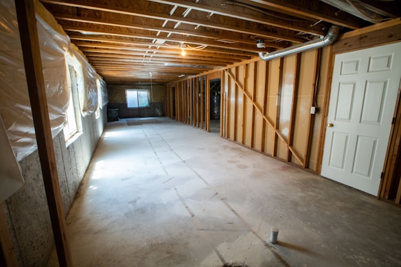 A spacious unfinished basement with exposed wooden beams on the ceiling and concrete floors. The walls are partially covered with wooden framing and plastic sheets. There is a door on the right side and a small window at the end of the room, providing some natural light. Pipes and ductwork are visible along the ceiling.
