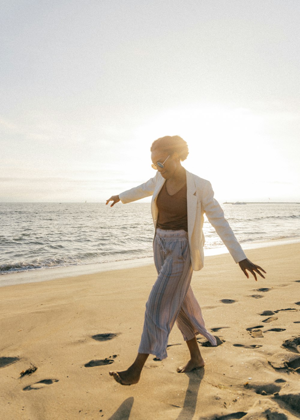 Frau in weißem Langarmhemd und grauer Hose geht tagsüber am Strand spazieren