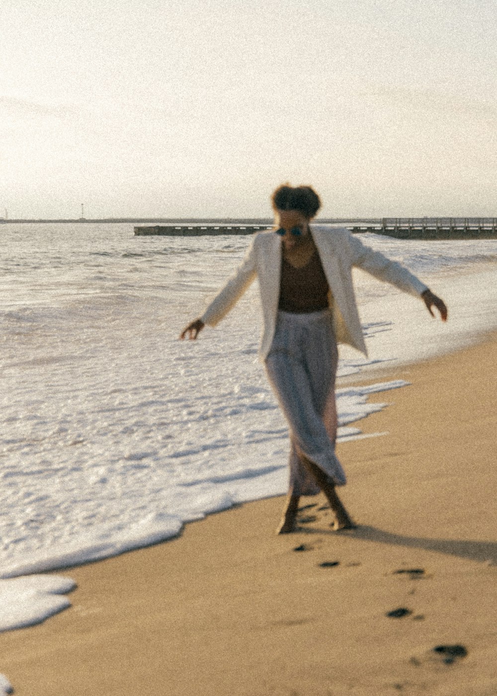 Frau in weißem Langarmhemd und grauer Hose geht tagsüber am Strand spazieren