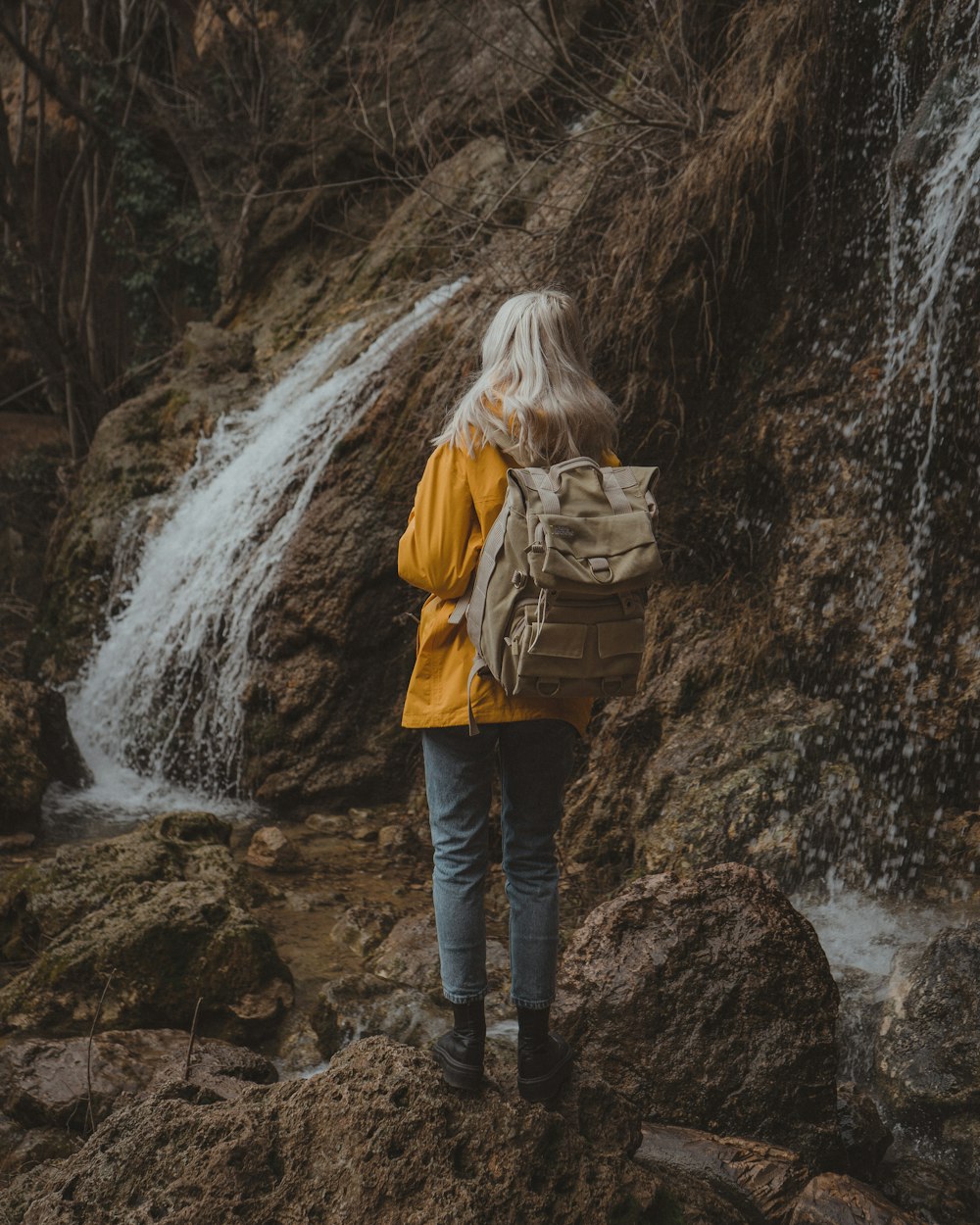 woman in brown jacket and blue denim jeans standing on rocky river during daytime