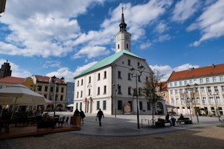 A town square featuring a large, historic building with a clock tower and a green roof. The square is surrounded by colorful buildings with red and yellow facades, and there are several street lamps and a few trees. People are casually walking or sitting on benches, and there's outdoor seating covered by umbrellas.