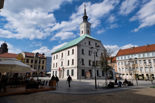 A town square featuring a large, historic building with a clock tower and a green roof. The square is surrounded by colorful buildings with red and yellow facades, and there are several street lamps and a few trees. People are casually walking or sitting on benches, and there's outdoor seating covered by umbrellas.