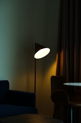 A dimly lit room with a modern floor lamp casting a warm glow. The lamp is adjacent to a dark-colored armchair and a round table. Heavy curtains add to the subdued ambiance.