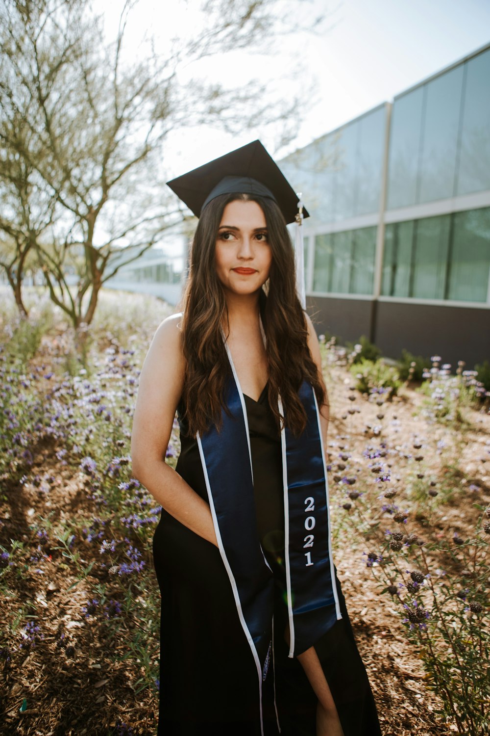 Femme en robe académique noire portant un chapeau académique noir debout près des arbres à feuilles vertes pendant la journée