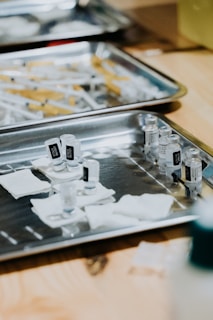 A medical setting with stainless steel trays containing multiple syringes and small vials, possibly of vaccine or medication. Gauze pads are also visible, suggesting preparedness for medical procedures.