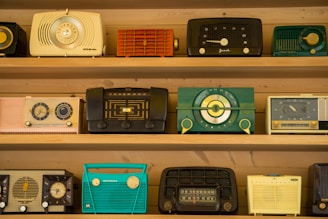 A collection of vintage radios is displayed on wooden shelves. Each radio has a distinct design and color, ranging from teal and black to yellow and red. The styles reflect a bygone era with dials, knobs, and mesh speaker coverings, showcasing the aesthetic diversity of mid-20th-century radio craftsmanship.