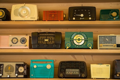 A collection of vintage radios is displayed on wooden shelves. Each radio has a distinct design and color, ranging from teal and black to yellow and red. The styles reflect a bygone era with dials, knobs, and mesh speaker coverings, showcasing the aesthetic diversity of mid-20th-century radio craftsmanship.